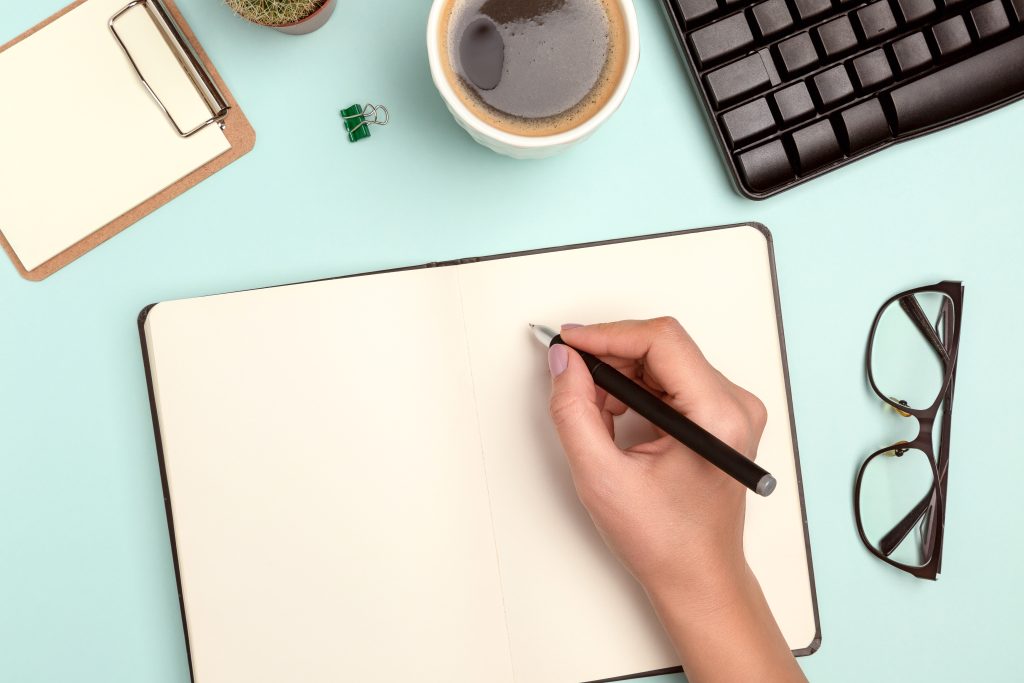 Women's hands hold a ballpoint pen. Coffee, laptop, notebook, cactus are at the workplace.